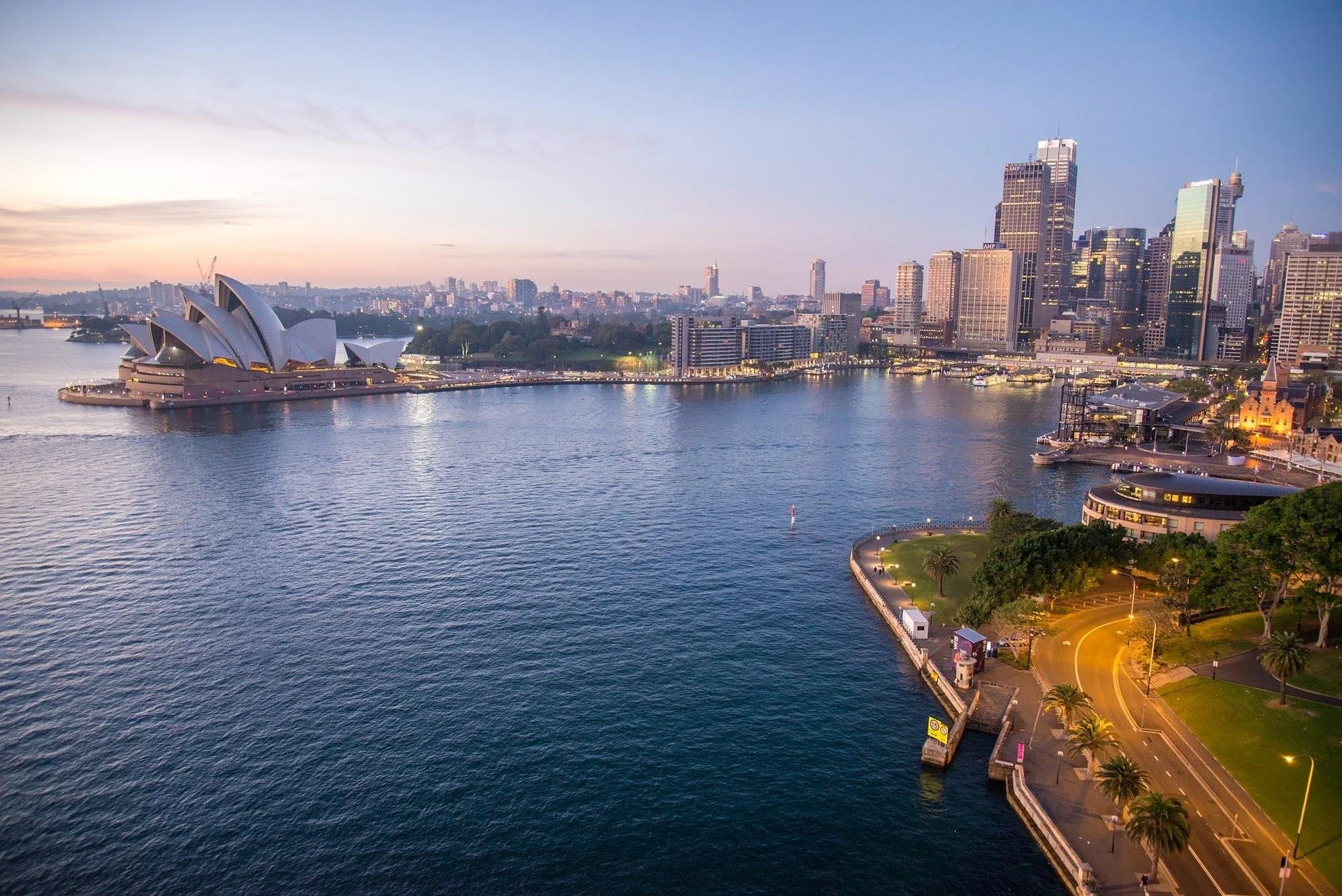 Aerial view of Sydney Harbour including the Sydney Opera House at sunset, with city buildings and water reflections.
