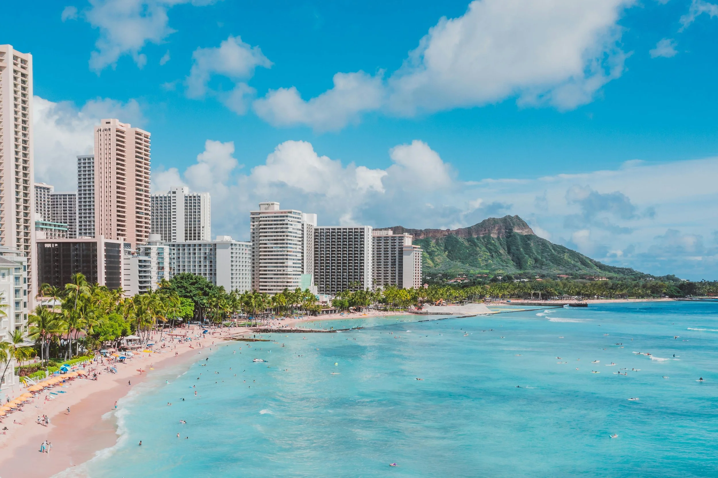 Beach with people, palm trees, and colorful umbrellas near high-rise buildings and a green mountain under a partly cloudy sky.