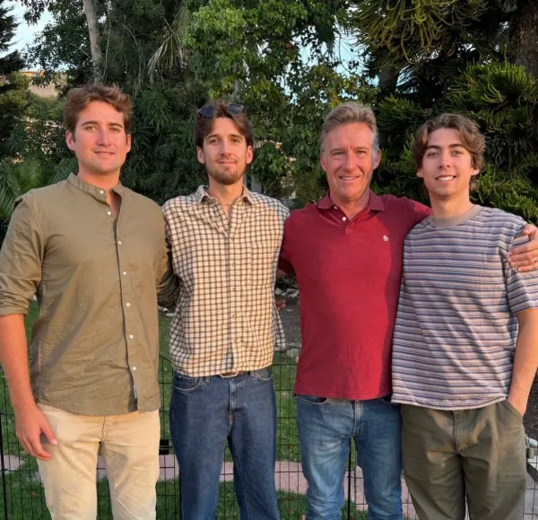 Four men standing outdoors in front of greenery, smiling, with their arms around each other's shoulders.